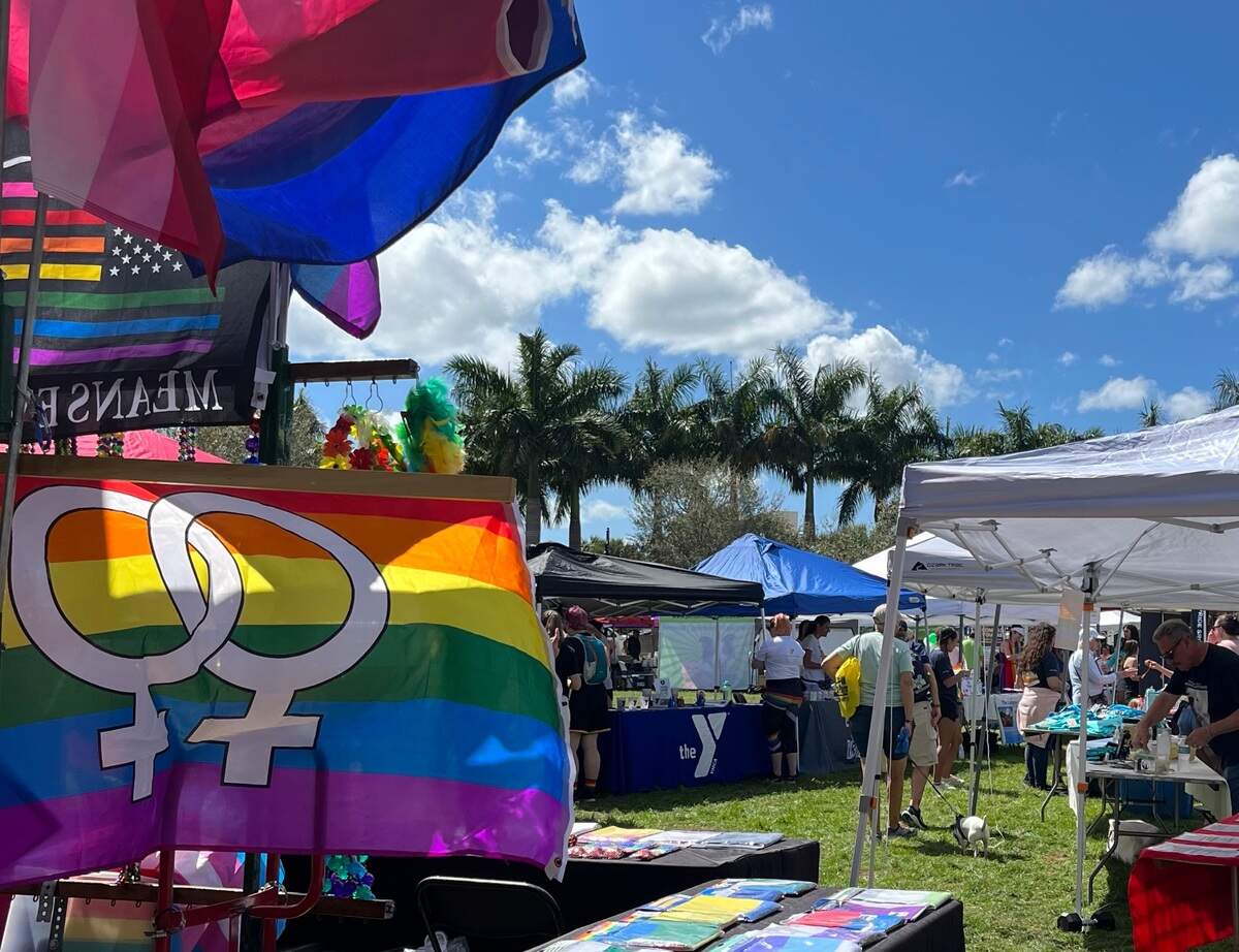 Rainbow-colored pride flag with double interlocking male symbols raised in the foreground, as guests browse vendors’ tables during the 7th annual Manatee County Pride festival at Riverwalk Park in March.