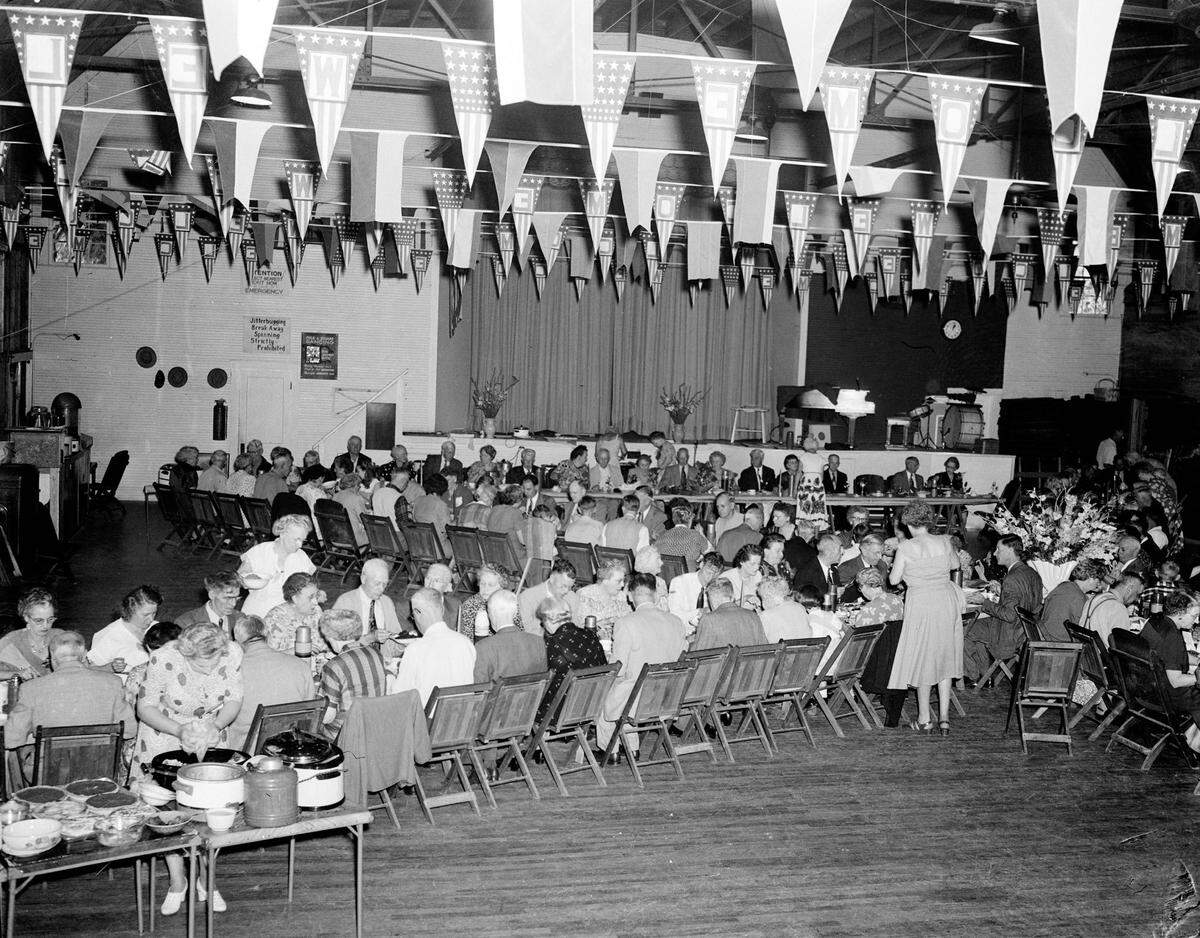 Residents dine in the auditorium at Bradenton Trailer Park owned by Kiwanis in 1951. The room is decorated with welcome flags and signs on the wall alert residents to emergency exits, warn that the Jitterbug dance, spinning and break away dance is strictly forbidden. Folk and square dancing events are advertised and dance band instruments are seen on stage. Photo from the Odom Photography Studio on Old Main Street in Bradenton.