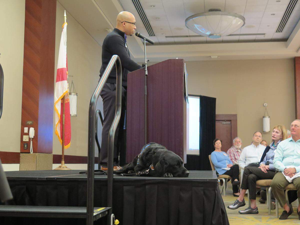 Pella, a black Labrador service dog from Southeastern Guide Dogs, lays on the stage near her veteran, Sean Brown while he speaks to a crowd gathered at the Southeastern Guide Dogs Heroes Breakfast on Wednesday at the Hyatt Regency Sarasota.