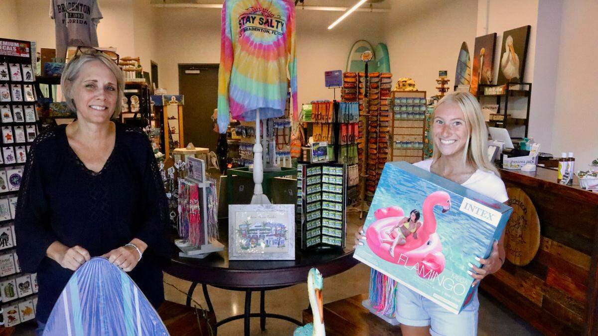 Sharon Hanzlik, left, shown 7/8/2022, holds a skim board, while Allison Bisker holds a pink flamingo float at The Beach Shop, which recently opened in the City Centre building on Bradenton’s Old Main Street.