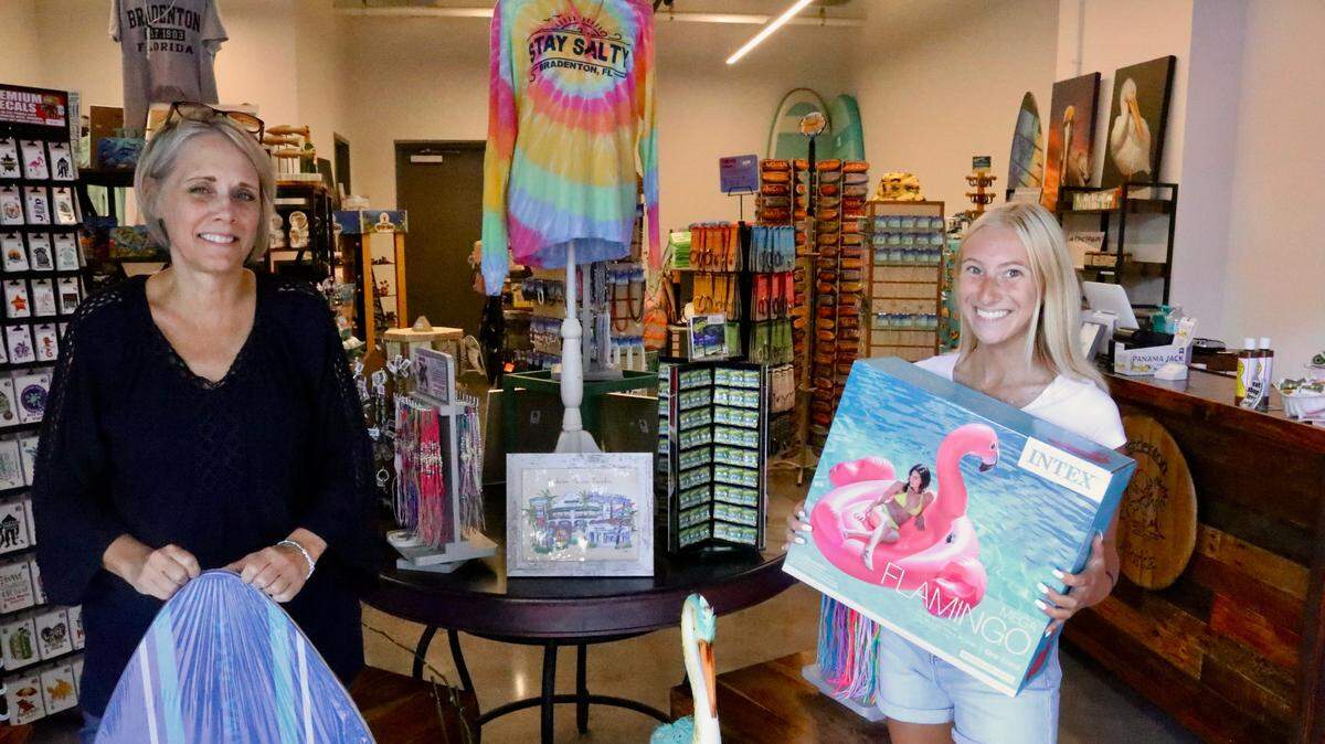 Sharon Hanzlik, left, shown 7/8/2022, holds a skim board, while Allison Bisker holds a pink flamingo float at The Beach Shop, which recently opened in the City Centre building on Bradenton’s Old Main Street.