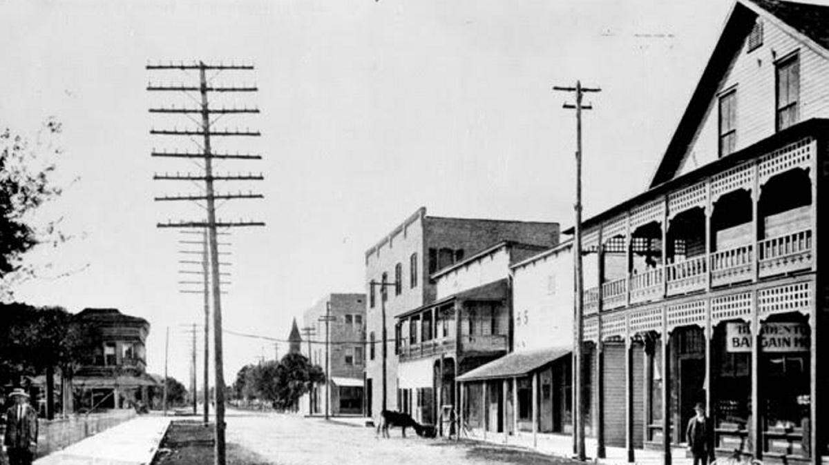 Looking west down Manatee Avenue from the corner of Broad Street (11th Street West) and Manatee Avenue. The Fuller building and facing building on Old Main Street are of brick, and there are nice paved sidewalks along Courthouse Square at left and in front of the stores at right, but the street is still unpaved and a free-range cow enjoys a nibble along the right side of the street. The buildings all have porches to guard against sun or rain. A gentleman stands near a propped-up bicycle and the main line of telephone poles stand along Manatee Avenue. 
 Manatee County Public Library Historic Image Digital collection