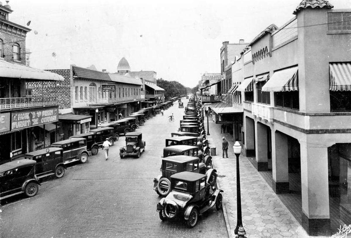 A view looking south along 12th Street West from 4th Avenue where multiple residents and visitors have parked their cars. Businesses like The Howard and Perry Furniture Company and the Fletcher Insurance Company line the east side of 12th street.