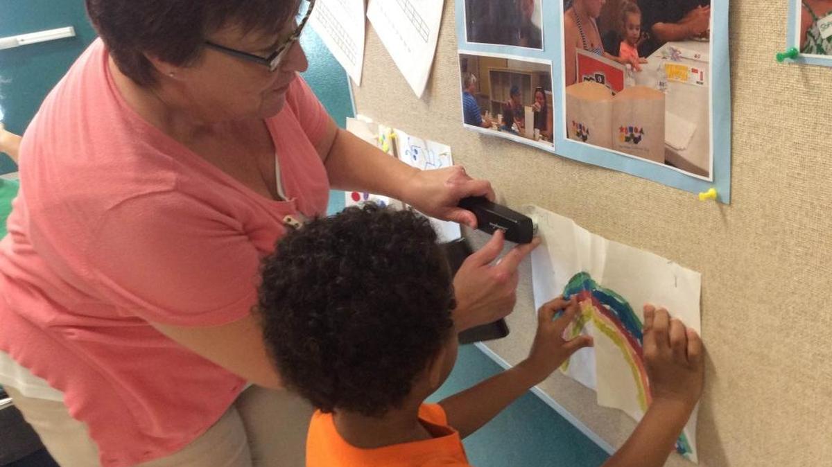 Luanne Howe, an early learning specialist with the Manatee County School District, helps 5-year-old David Weiland staple a homework project to a bulletin board at Sea Breeze Elementary School.