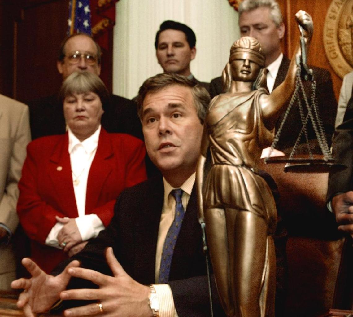 Behind the sculpture of Justice on his Capitol desk, Gov. Jeb Bush comments while defending his approval of a gun bill preventing local governments from suing gun manufacturers after he signed the measure into law May 1, 2001. NRA lobbyist Marion Hammer is in the red jacket at left.